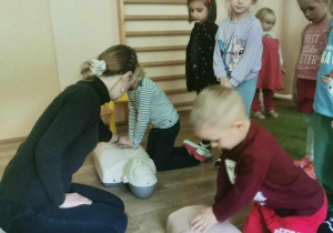 Prowadząca zajęcia obserwuje dwoje dzieci ćwiczące usiskanie klatki piersiowej przy resuscytacji (na fantomach).