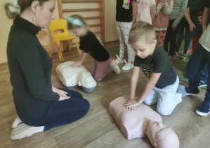 Prowadząca zajęcia obserwuje dwoje dzieci ćwiczące usiskanie klatki piersiowej przy resuscytacji (na fantomach).