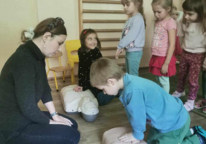 Prowadząca zajęcia obserwuje dwoje dzieci ćwiczące usiskanie klatki piersiowej przy resuscytacji (na fantomach).
