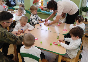 Dzieci z najmłodszej grupy wraz z nauczycielkami i żołnierzem przy stoliku wykonują kotyliony.