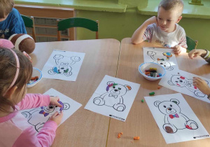 Dzieci siedzą przy stoliku wyklejając plasteliną kontur misia.