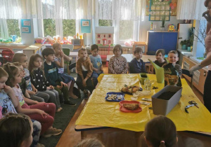Dzieci słuchają wypowiedzi nauczycielki, siedzą na krzesełkach wokół stołu, na którym są różne przybory ogrodnicze.