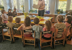 Dzieci słuchają wypowiedzi nauczycielki, siedzą na krzesełkach wokół stołu, na którym są różne przybory ogrodnicze.