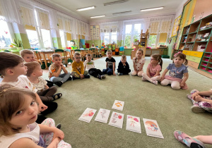 Dzieci siedzą na dywanie, na którym leżą obrazki tematycznie związane z Świętami Wielkanocnymi.