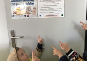 Dzieci wskazujące palcami plakat promujący zbieranie plastikowych butelek.