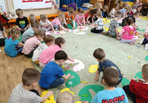 Grupa dzieci siedzi na dywanie, wykleja skrzydła kolorowymi piórkami.
