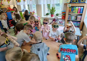 Dzieci stoją przy stoliku i prezentują się w okularach przeciwsłonecznych.