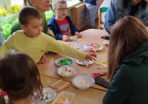 Grupa rodziców i dzieci wykonujących ozdoby wielkanocne przy stolikach.