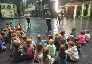 Grupa dzieci siedzacych na podłodze sceny Teatru Wielkiego, słucha opowieści Pani oprowadzajacej po teatrze.