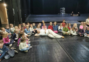 Grupa dzieci siedzacych na podłodze sceny Teatru Wielkiego pozuje do zdjęcia wraz z odtwórczynia roli Arielki.