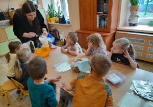 Dzieci siedzą przy stoliku i obserwują jakie produkty pani wsypuje do miseczki.
