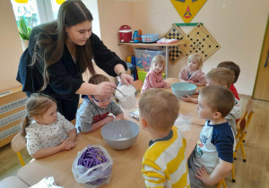 Dzieci siedzą przy stoliku i obserwują jakie produkty pani wsypuje do miseczki.