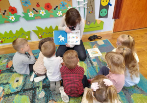 Grupa dzieci siedzi na dywanie i słucha czytaną przez panią książkę.