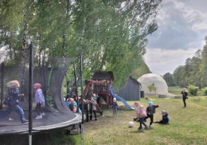 Dzieci bawią się na placu zabaw. Po lewej stronie stoi trampolina, na której skaczą trzy dziewczynki. W tle widać namiot sferyczny.