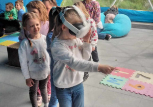 Dziewczynki stoja w kolejce. Dziewczynka na początku ma założone gogle VR i wskazuje palcem, gdzie widzi dinozaura.