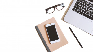 Fotografia przedstawiająca leżące: laptop, telefon, notes, długopis i okulary.